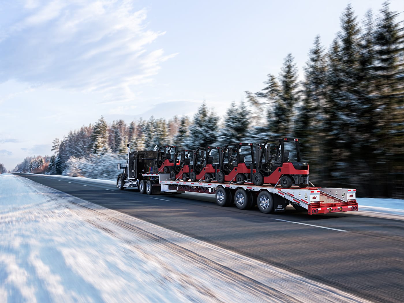 a flat bed trailer carrying fork lifts drives down the road between snow-covered trees